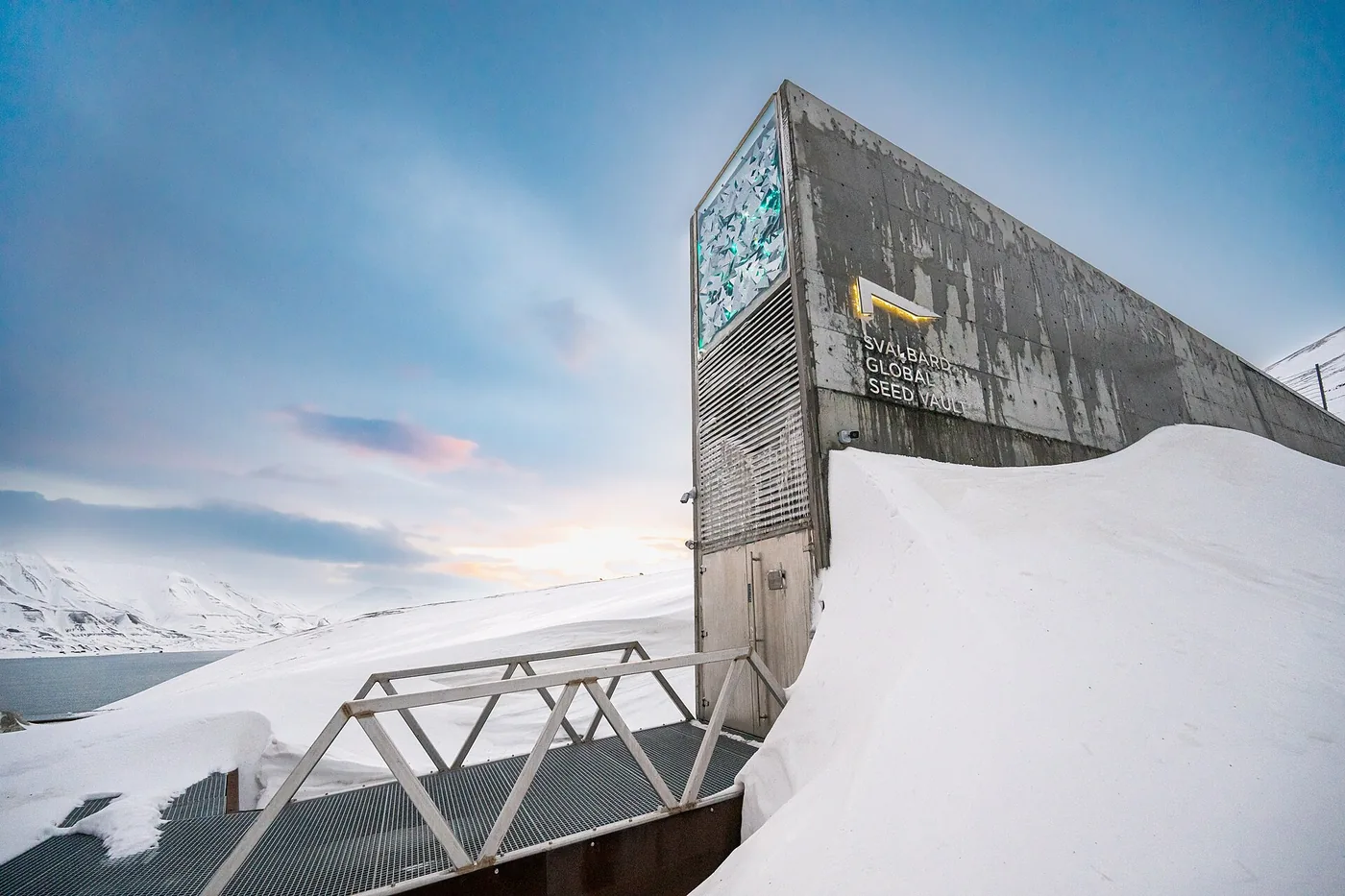 Fachada de concreto da entrada do cofre de sementes de Svalbard, cercada de neve, com uma passarela metálica à frente.
